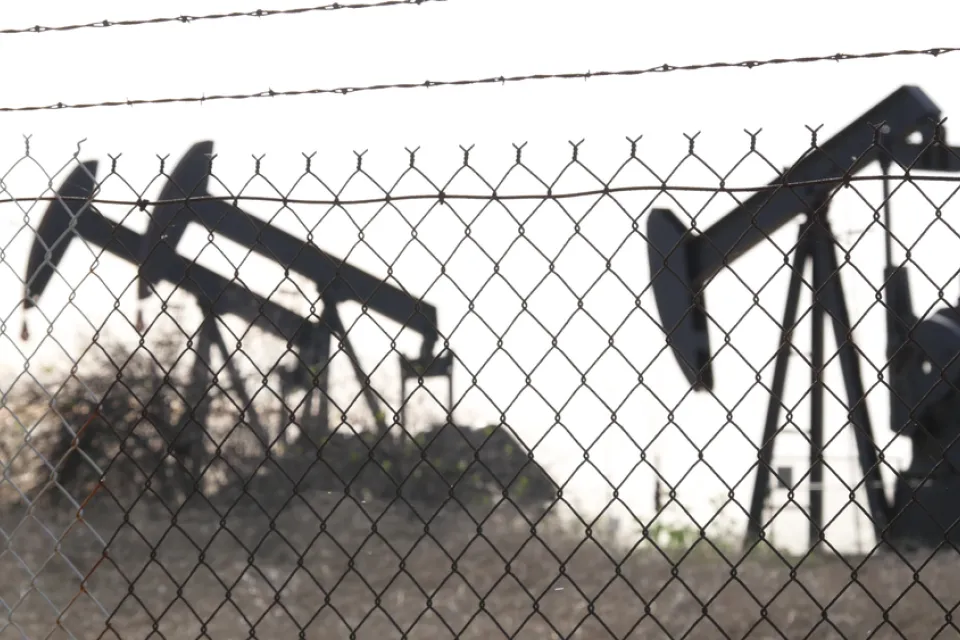 View of black oil wells behind chain link fence with barbed wire top